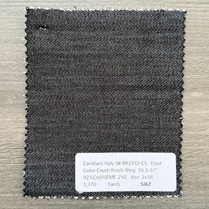 May include: A rectangular swatch of dark denim fabric with a white selvage edge. The fabric has a textured appearance. A white label with black text provides details about the fabric, including the name, composition, and measurements.