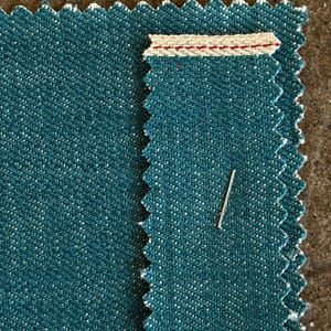 May include: Close-up of two teal fabric swatches with a zigzag edge. One swatch has a cream-colored band with red stitching. A silver pin is inserted into the fabric. The fabric has a speckled texture.