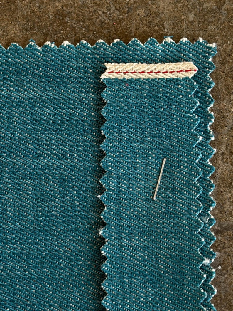 May include: Close-up of two teal fabric swatches with a zigzag edge. One swatch has a cream-colored band with red stitching. A silver pin is inserted into the fabric. The fabric has a speckled texture.