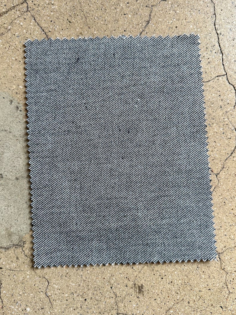 May include: A close-up of a gray denim fabric swatch. The fabric has a subtle diagonal weave pattern.