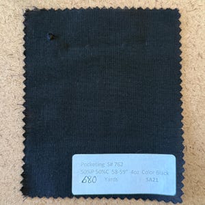 May include: A black fabric swatch with a jagged edge. A white label displays text: "Pocketing S# 762 50%P 50%C 58-59" 4oz Color Black 680 Yards SA21". The fabric appears to be a sample of a pocketing material.
