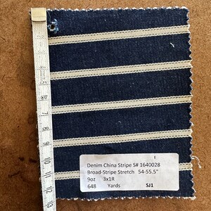 Puede incluir: Una muestra de tejido vaquero con rayas blancas sobre un fondo azul oscuro. El tejido est&aacute; etiquetado como "Denim China Stripe S# 1640028 Broad-Stripe Stretch 54-55.5" 9oz 3x1R Yards SJ1 648"