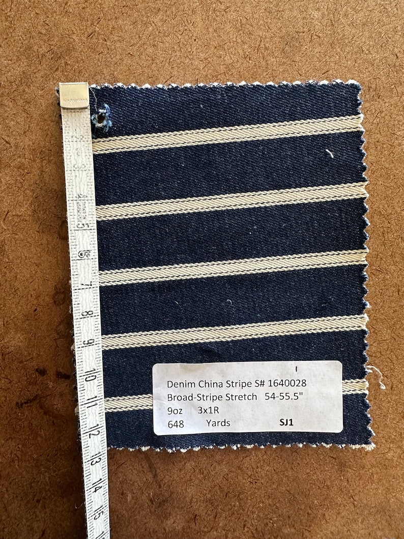 Puede incluir: Una muestra de tejido vaquero con rayas blancas sobre un fondo azul oscuro. El tejido est&aacute; etiquetado como "Denim China Stripe S# 1640028 Broad-Stripe Stretch 54-55.5" 9oz 3x1R Yards SJ1 648"