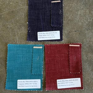 May include: Three fabric swatches in shades of purple, turquoise, and red. Each swatch has a white label with text indicating the fabric type, color, and measurements of 39.5 inches and 13.5 ounces. The fabrics are labeled as Purple, Turquoise, and Red Selvedge Denim.