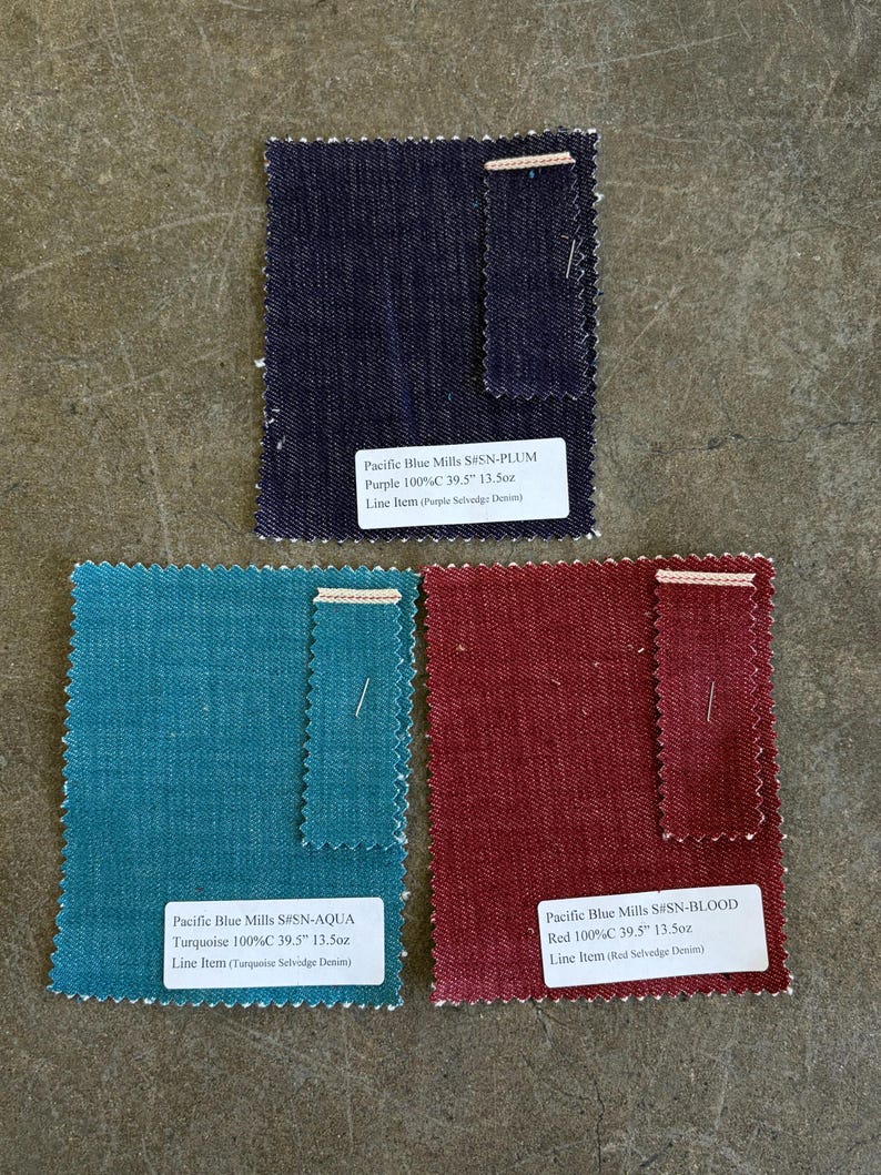 May include: Three fabric swatches in shades of purple, turquoise, and red. Each swatch has a white label with text indicating the fabric type, color, and measurements of 39.5 inches and 13.5 ounces. The fabrics are labeled as Purple, Turquoise, and Red Selvedge Denim.