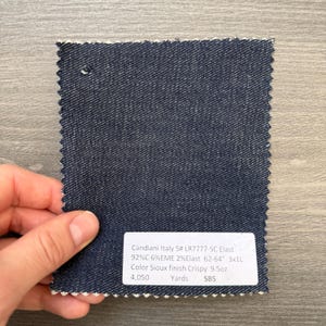 May include: A square denim fabric swatch with a white label. The fabric is a dark blue with a textured appearance. The label reads "Candiani Italy S# LR7777-SC Elast" and other details about the fabric composition and weight.