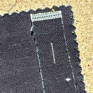 May include: Close-up of a dark blue denim fabric with a white stitching detail. A small pin is inserted into the fabric.