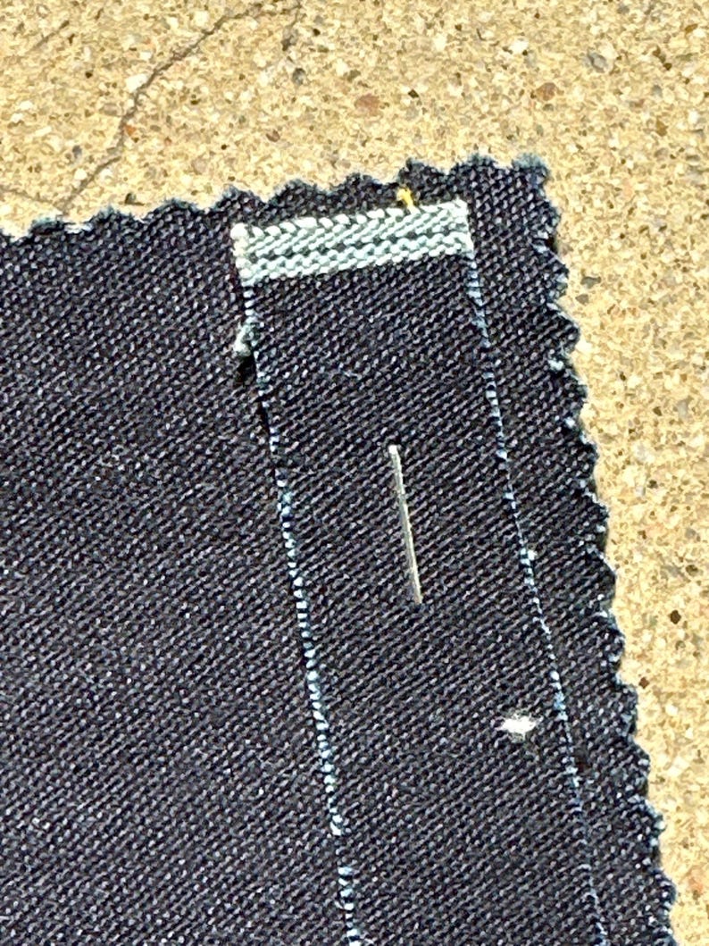 May include: Close-up of a dark blue denim fabric with a white stitching detail. A small pin is inserted into the fabric.