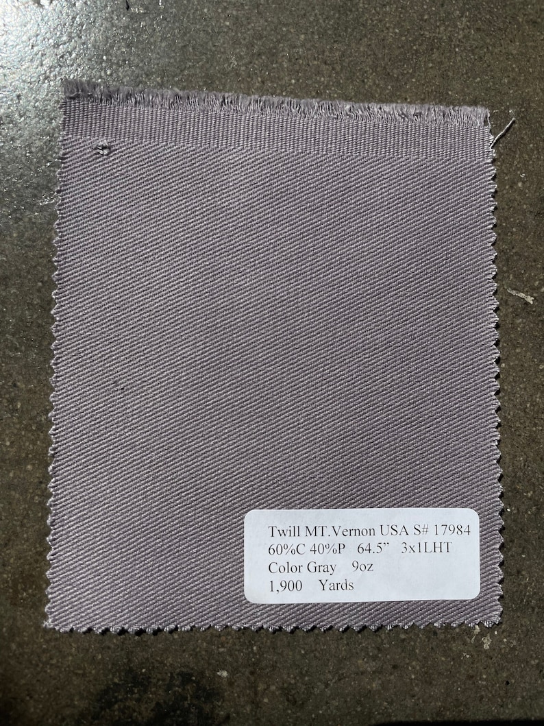 Op de afbeelding: Een close-up van een grijze twill stofstaal met een wit label met de tekst "Twill MT.Vernon USA S# 17984 60%C 40%P 64.5" 3x1LHT Kleur Grijs 9oz 1.900 Yards".