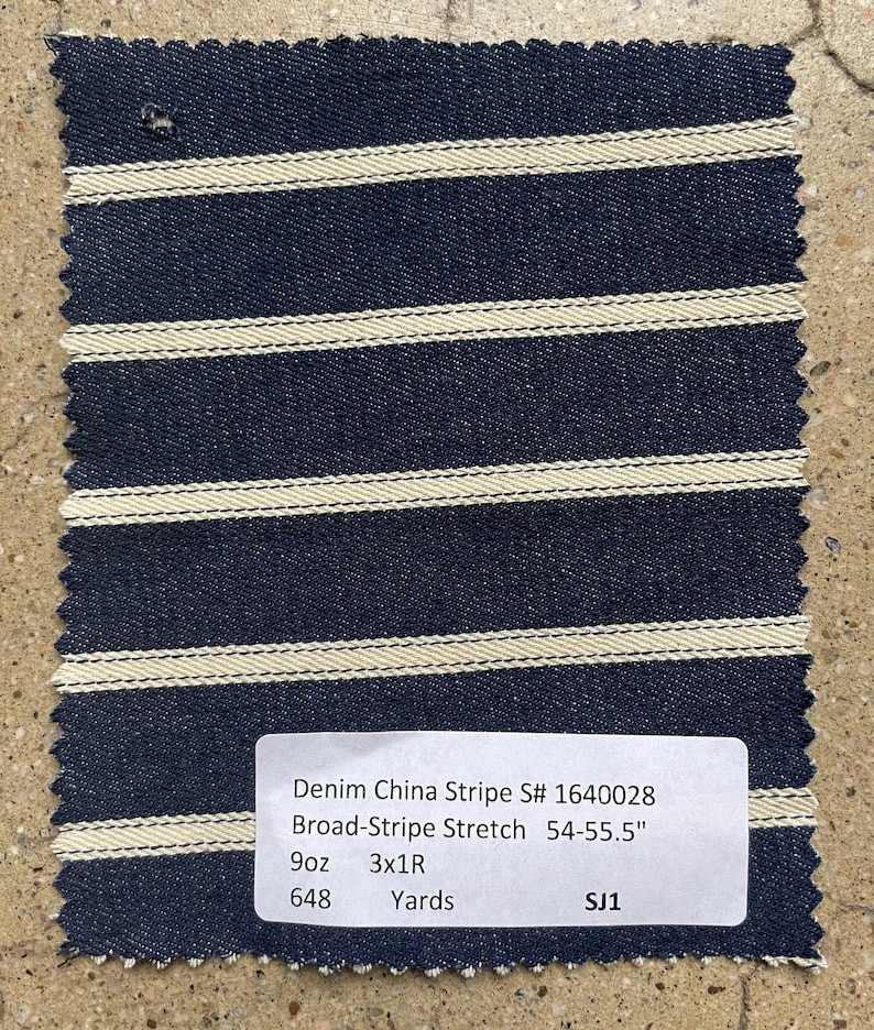 Puede incluir: Un primer plano de una muestra de tela a rayas azul marino y blanco con una etiqueta que dice "Denim China Stripe S# 1640028 Broad-Stripe Stretch 54-55.5" 9oz 3x1R 648 Yards SJ1".
