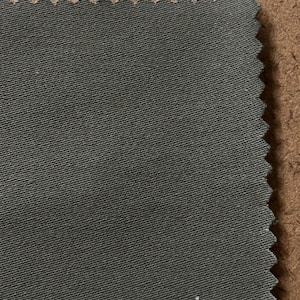 May include: A swatch of dark gray fabric with a textured surface and a zigzag edge. The fabric is set against a brown background, showcasing its color and texture. Ideal for textile projects.