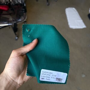 May include: A green fabric swatch with a white label that reads "Twill S# LB16 65%P 35%C 57-58" Color Green 4.7oz 1000 Yards YARD".