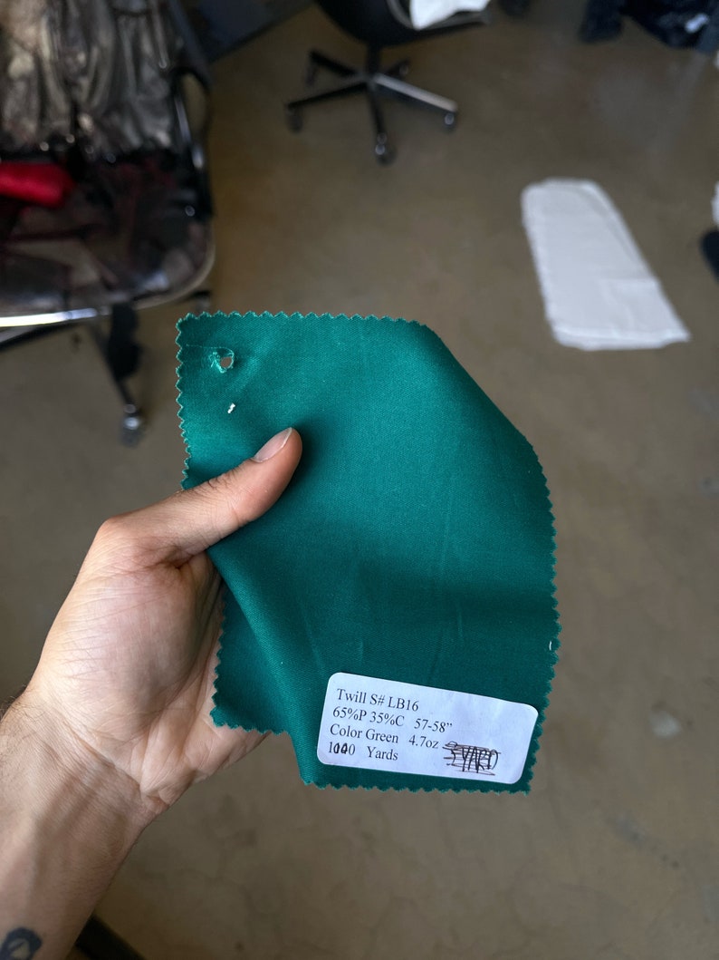 May include: A green fabric swatch with a white label that reads "Twill S# LB16 65%P 35%C 57-58" Color Green 4.7oz 1000 Yards YARD".