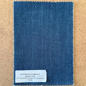 May include: A rectangular swatch of dark blue denim fabric with a white selvage edge. The fabric has a textured appearance. A white label at the bottom reads "KAIHARA 6720 BRW ACT Broken Twill 57" 12 oz 98% Cotton 2% Spandex Yards."
