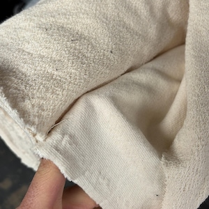 Może przedstawiać: Zbliżenie na rolkę kremowej tkaniny. Tkanina wydaje się być materiałem o dużej gramaturze, prawdopodobnie frotte lub podobnym chłonnym materiale. Widoczna jest faktura, z pętelkową powierzchnią po jednej stronie i gładszą, płaską powierzchnią po drugiej.