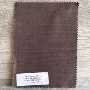 May include: A rectangular swatch of brown fabric with a textured surface and a zig-zag edge. A white label displays the text "Mt. Vernon 26034 Lt. Brown Ripstop 66" 7 oz 100% Cotton yards."