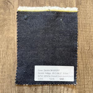 May include: A rectangular swatch of dark blue denim fabric with a frayed edge and a yellow stripe. A white label with black text provides details about the fabric, including the name, weight, and composition.