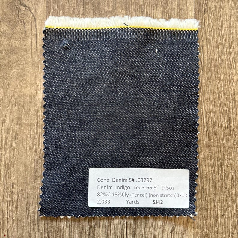 May include: A rectangular swatch of dark blue denim fabric with a frayed edge and a yellow stripe. A white label with black text provides details about the fabric, including the name, weight, and composition.