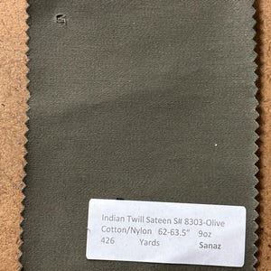 May include: Olive green Indian Twill Sateen fabric swatch. The fabric is Cotton/Nylon blend, 62-63.5 inches wide, and 9oz. A white label displays the fabric details: "Indian Twill Sateen S# 8303-Olive", "426 Yards", and "Sanaz".