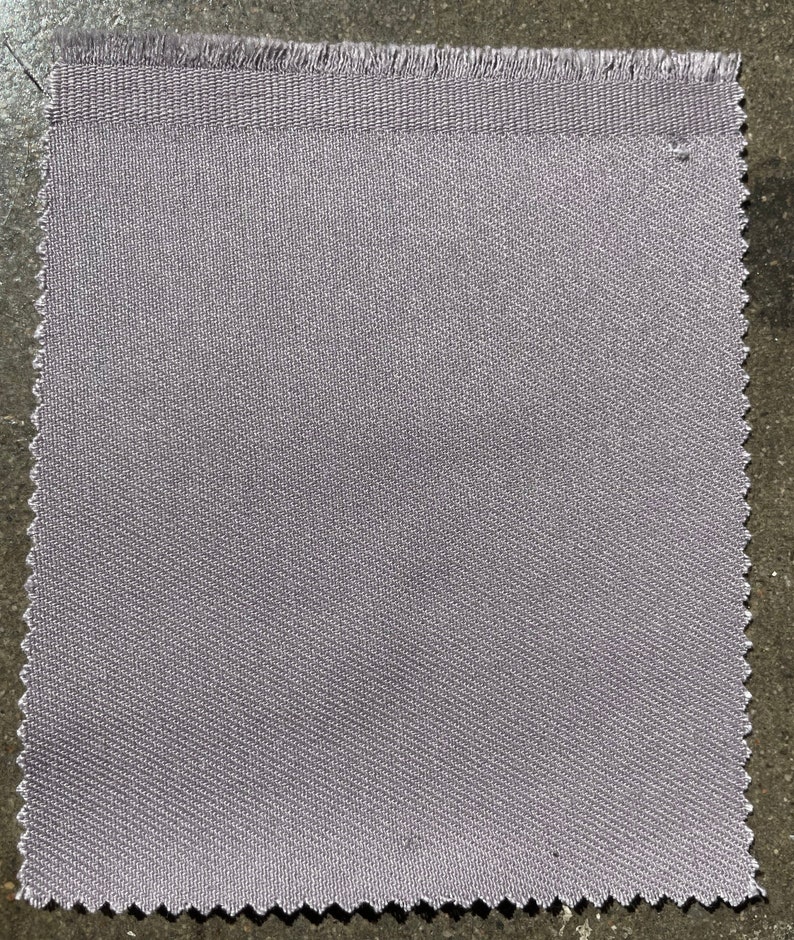 Op de afbeelding: Een close-up van een stuk lichtgrijs stof met een textuurweefsel. De stof heeft een rauwe rand.