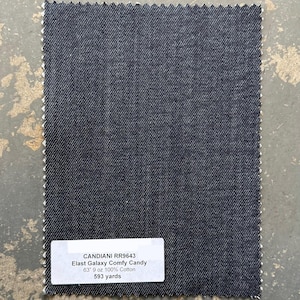 May include: A rectangular swatch of dark blue denim fabric with a textured surface. The fabric has a white, zig-zag edge. A white label at the bottom displays text including "CANDIANI RR9643" and "Elast Galaxy Comfy Candy".