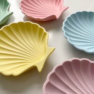 May include: Five seashell-shaped dishes in different colours: light blue, light pink, yellow, green, and a darker pink. The dishes are made of a smooth, textured material and are perfect for serving small snacks or desserts.