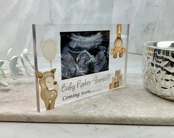 Könnte beinhalten: Ein klarer Acryl-Bilderrahmen mit weißem Hintergrund. Der Rahmen enthält ein Schwarzweiß-Ultraschallbild und eine handschriftliche Nachricht mit dem Text "Baby Parker-James Coming Soon..." mit einem Herzsymbol. Der Rahmen zeigt außerdem eine kleine Hirschfigur mit einem Ballon und eine Giraffenfigur.