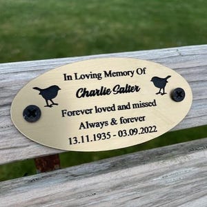 May include: Gold oval memorial plaque with black engraved text: "In Loving Memory Of Charlie Salter Forever loved and missed Always & forever 13.11.1935 - 03.09.2022". Two black metal screws are attached to the plaque. Two black silhouettes of birds are engraved on the plaque.