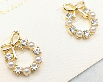 May include: A pair of gold-tone earrings with a wreath design. Each earring features a small bow at the top and is adorned with clear rhinestones and white pearls.