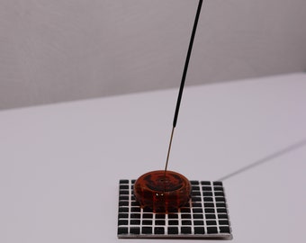 May include: A decorative incense holder featuring a dark amber resin disc with a swirling pattern, set on a black and white tiled base. A black incense stick is inserted into the resin. The holder is designed for burning incense.