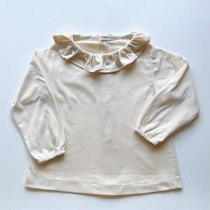 May include: A cream-coloured long-sleeved shirt with a ruffled collar. The shirt has a relaxed fit and is made of a soft, comfortable fabric.