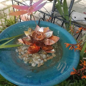 May include: A blue ceramic bird bath with a copper lotus flower and dragonfly sculpture. The water in the bath is clear and there are small white rocks at the bottom.