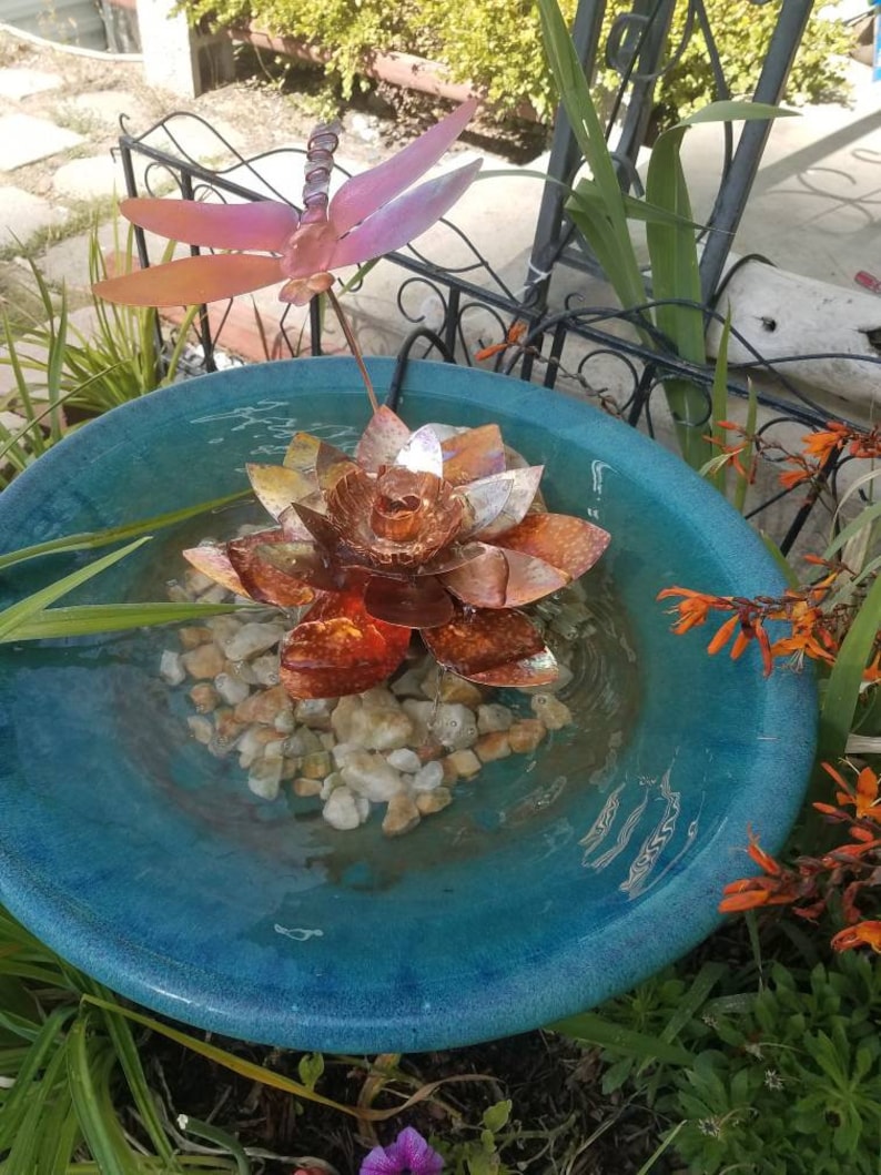 May include: A blue ceramic bird bath with a copper lotus flower and dragonfly sculpture. The water in the bath is clear and there are small white rocks at the bottom.