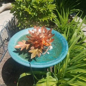 以下が含まれることがあります： 蓮の花のデザインが施された銅製のバードバスの噴水。噴水は緑の植物に囲まれ、青い台座の上に置かれています。