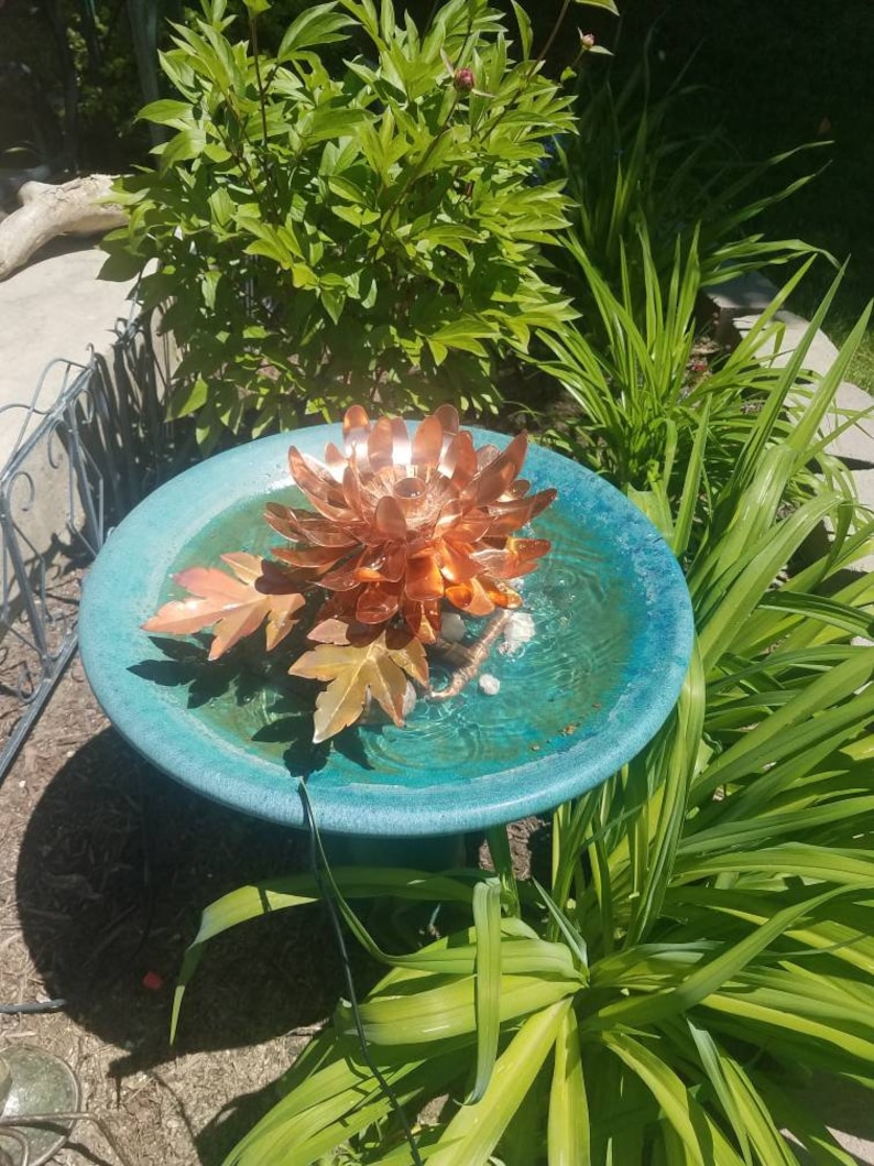 以下が含まれることがあります： 蓮の花のデザインが施された銅製のバードバスの噴水。噴水は緑の植物に囲まれ、青い台座の上に置かれています。
