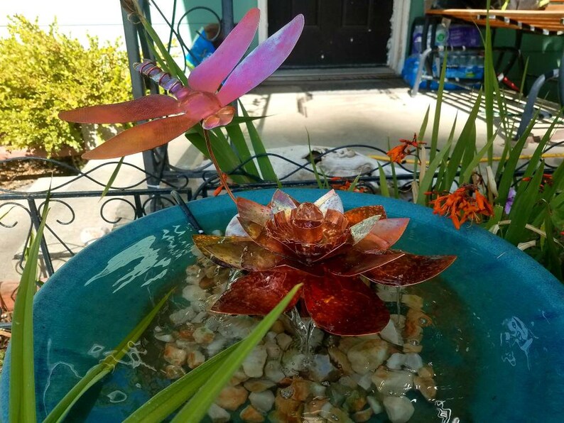 May include: A copper-colored metal lotus flower fountain with a dragonfly sculpture above it. The fountain is in a blue ceramic bowl with water and rocks.