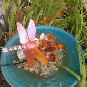 May include: A teal blue ceramic bird bath with a copper dragonfly sculpture in the center. The dragonfly has pink and orange wings and is surrounded by small white rocks.