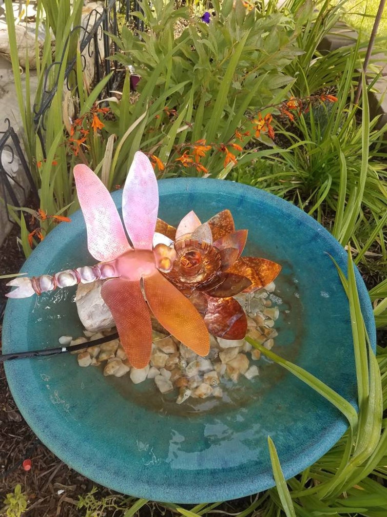 May include: A teal blue ceramic bird bath with a copper dragonfly sculpture in the center. The dragonfly has pink and orange wings and is surrounded by small white rocks.
