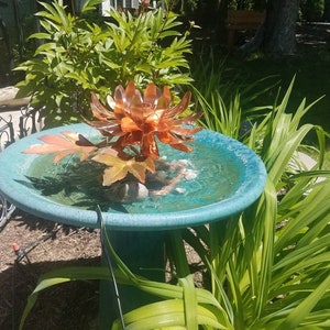 以下が含まれることがあります： 中央に銅の花模様が施された銅製のバードバス。バードバスは青い台座の上に置かれ、緑の植物に囲まれています。