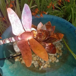 May include: A copper dragonfly sculpture with iridescent wings sits atop a copper flower in a blue birdbath. The birdbath is filled with water and small stones.