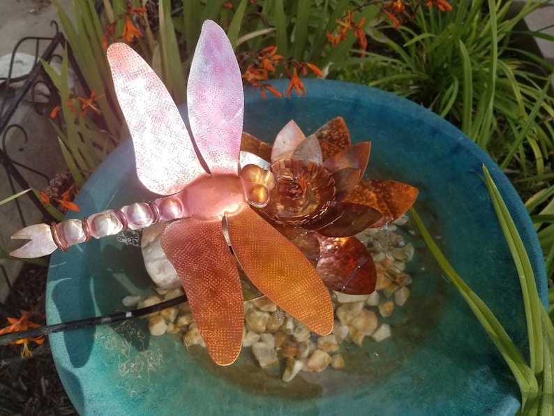 May include: A copper dragonfly sculpture with iridescent wings sits atop a copper flower in a blue birdbath. The birdbath is filled with water and small stones.