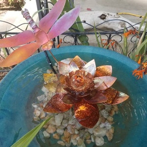 May include: A copper-colored metal lotus flower fountain with a dragonfly sculpture on a metal rod. The fountain is in a blue ceramic bowl with water and small rocks.