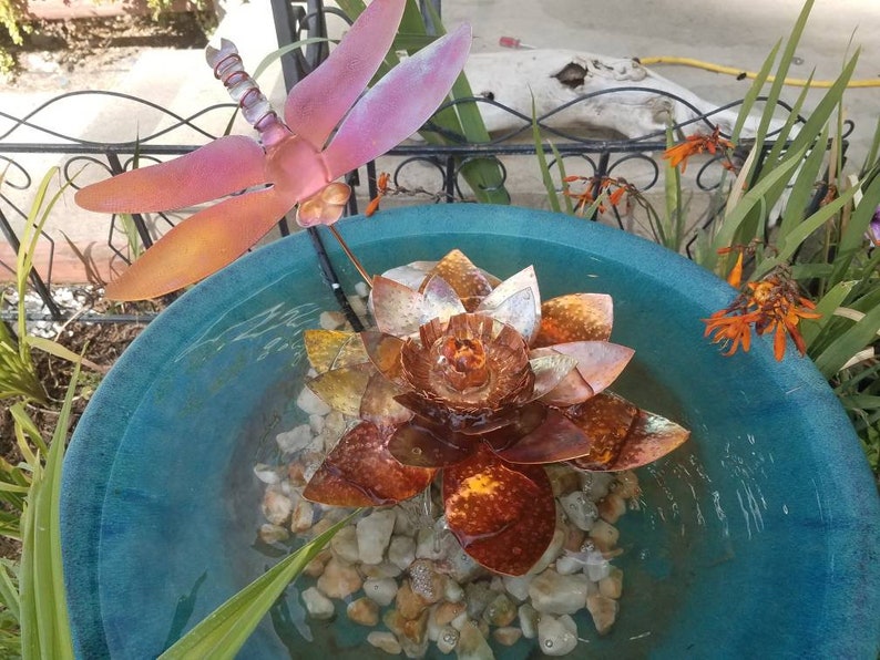 May include: A copper-colored metal lotus flower fountain with a dragonfly sculpture on a metal rod. The fountain is in a blue ceramic bowl with water and small rocks.