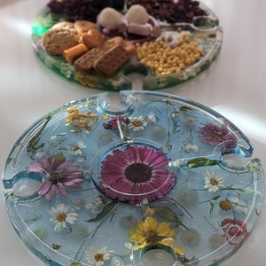 Könnte beinhalten: Ein rundes, klares Serviertablett aus Harz mit blauem Hintergrund und gepressten Blumen und Blättern. Das Tablett hat vier Aussparungen zum Halten von Gläsern.