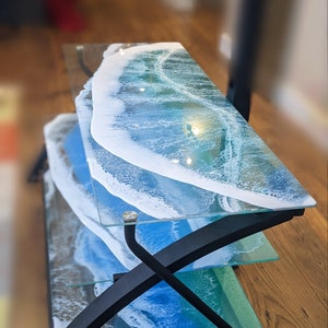 May include: A three-tiered glass and metal stand with a wave design. The glass shelves feature a blue and white resin pour with white swirls resembling ocean waves. The stand has a black metal frame with a curved design.