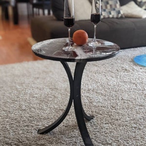Op de afbeelding: Een kleine, ronde bijzettafel met een donker, patroonblad en zwarte, gebogen poten. Twee champagneglazen gevuld met rode wijn en een sinaasappel staan op de tafel. De tafel staat op een beige tapijt.