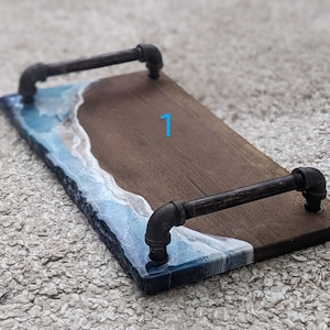 Peut inclure: Un plateau de service en bois avec un design d'onde océanique en résine bleue et blanche. Le plateau a deux poignées en métal noir.