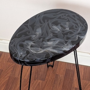 Op de afbeelding: Zwarte ovale bijzettafel met een wit marmerachtig harsblad en zwarte haarspeldpoten.