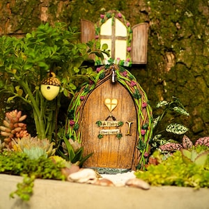 May include: A miniature fairy door with a wooden door and a heart-shaped cutout. The door is surrounded by green vines with pink flowers. The door has a sign that reads "A Fairy Elves Home".