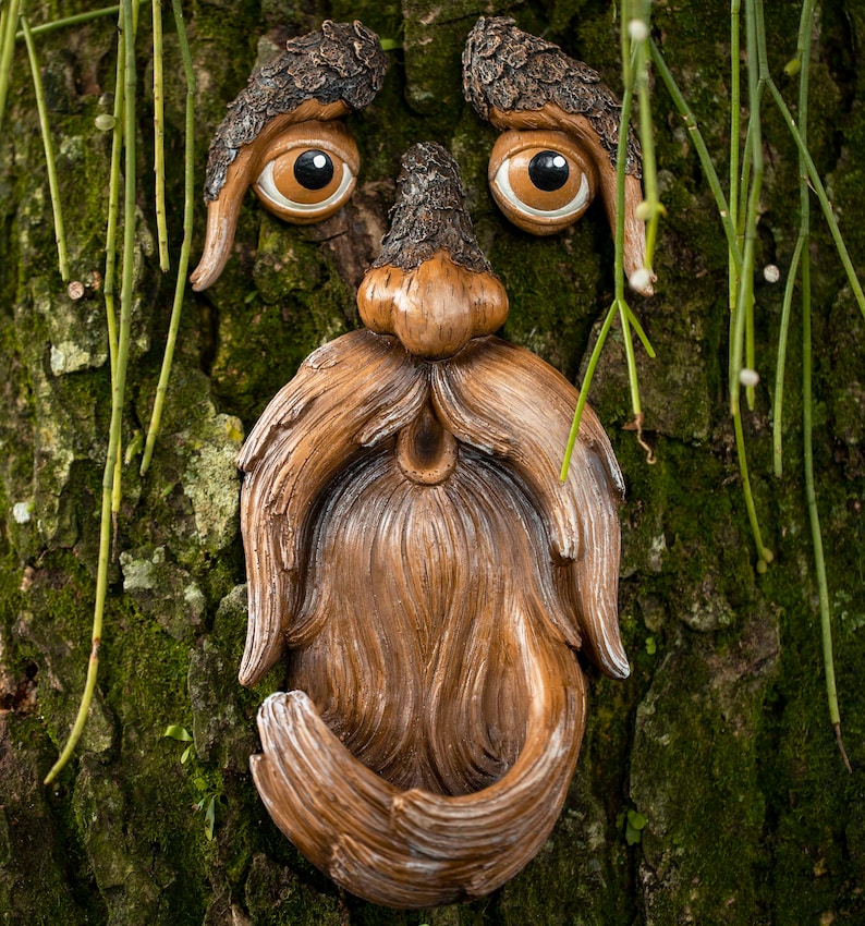 Puede incluir: Una escultura caprichosa de una cara de &aacute;rbol con ojos grandes y expresivos, una nariz y una larga barba fluida. La escultura est&aacute; hecha de un material marr&oacute;n y est&aacute; montada en un tronco de &aacute;rbol cubierto de musgo verde.